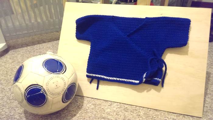Blue crochet baby kimono with white hem stripe, pictured next to a size 5 white and blue soccer ball